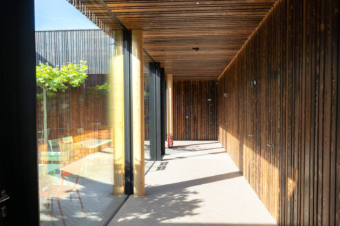 Überdachter Gang mit Glasfront und Holzverkleidung, der auf eine möblierte Außenterrasse mit bepflanzten Bäumen führt.