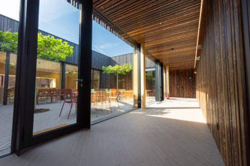 Heller Korridor mit Holzdecken und Glaswänden, der einen Übergang von innen nach außen schafft, mit Sicht auf bunte Außenmöbel.