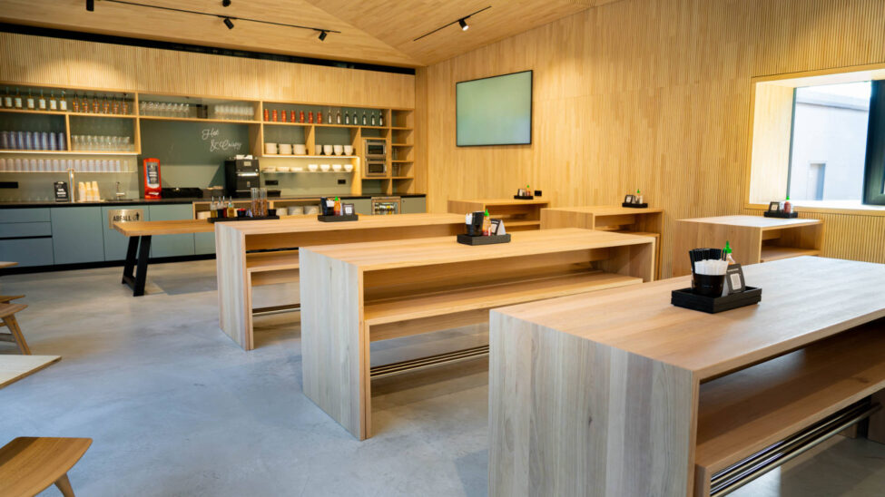 Offener Gastronomiebereich mit minimalistischen Holztischen und gut organisiertem Selbstbedienungstresen im Hintergrund.