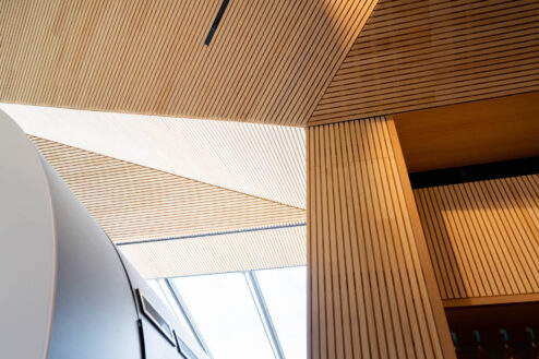 Architektonisches Detailbild der Decke mit schräg verlaufenden Holzlamellen und natürlichem Lichteinfall durch ein Oberlicht.