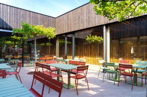 Farbenfrohe Außenterrasse mit roten und grünen Stühlen und Tischen, eingerahmt von dunkler Holzarchitektur und bepflanzten Bäumen.