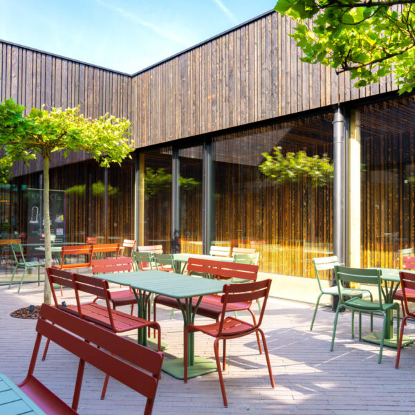 Farbenfrohe Außenterrasse mit roten und grünen Stühlen und Tischen, eingerahmt von dunkler Holzarchitektur und bepflanzten Bäumen.