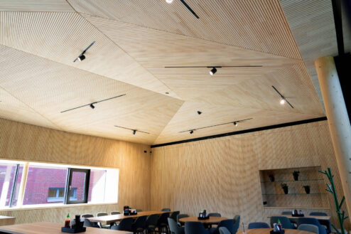 Innenraum mit geometrischer Holzdecke, großen Fenstern und modernem Sitzbereich mit runden Tischen und bequemen Stühlen.