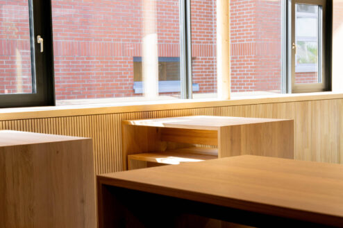 Sitzbereich am Fenster mit klarer Holzverkleidung und Blick auf eine rote Backsteinfassade im Außenbereich.