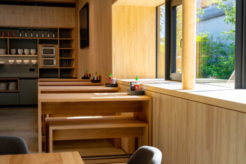 Reihen aus schlichten Holztischen an einem sonnendurchfluteten Fenster mit Blick auf grüne Außenflächen.