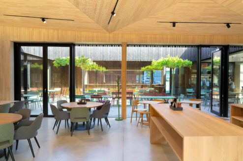 Innenraum mit großem Glasfenster und Blick in den begrünten Innenhof mit bunten Stühlen und Bäumen.