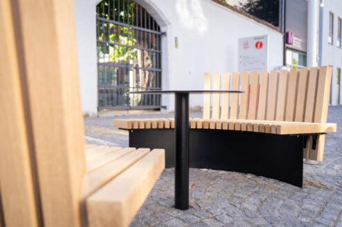 Runde Sitzbank aus hellem Holz mit schwarzem Metalltisch in der Mitte auf Kopfsteinpflaster.