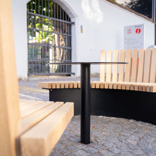 Runde Sitzbank aus hellem Holz mit schwarzem Metalltisch in der Mitte auf Kopfsteinpflaster.