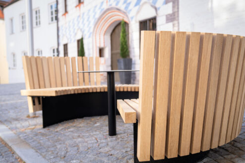 Zwei geschwungene Holzsitzbänke mit hoher Rückenlehne und zentralem Tisch vor dem Museum Dingolfing.
