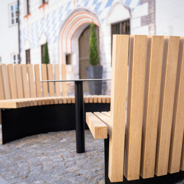 Zwei geschwungene Holzsitzbänke mit hoher Rückenlehne und zentralem Tisch vor dem Museum Dingolfing.