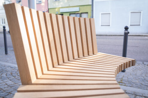Detailaufnahme einer gebogenen Holzbank mit schräg angeordneten Leisten und ergonomischem Design.