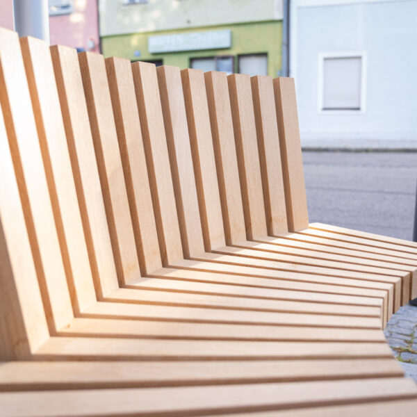Detailaufnahme einer gebogenen Holzbank mit schräg angeordneten Leisten und ergonomischem Design.